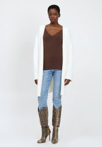 Langer weißer Cardigan über einem braunen V-Ausschnitt-Pullover, kombiniert mit hellblauen Jeans und kniehohen, gemusterten Schlangenlederstiefeln.