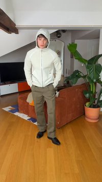 Jeune homme portant une veste à capuche blanche, un pantalon vert olive et des chaussures noires se tient dans un salon avec un sol en bois et une grande plante verte en pot.