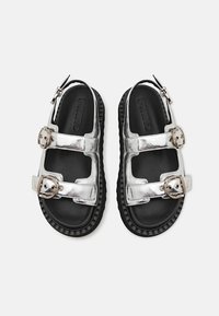 Ασημένια μεταλλικά σανδάλια με δύο φαρδιές λωρίδες, ρυθμιζόμενες αγκράφες και μαύρη αυλακωτή καουτσούκ σόλα. Λείο υλικό με μινιμαλιστική σχεδίαση.