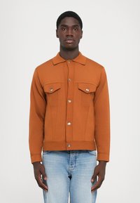 Chaqueta marrón con textura y cierre de botones en la parte delantera, dos bolsillos en el pecho y detalles en plata. Combinada con jeans de color azul claro.