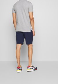Man som bär en grå kortärmad skjorta, marinblå shorts och färgglada Polo-sneakers, står vänd mot en enkel ljusgrå vägg.
