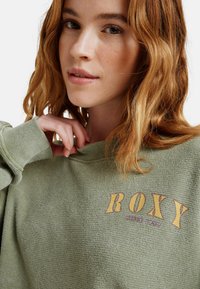 Jonge vrouw met golvend rood haar in een groene sweater met textuur en gele "ROXY"-tekst, terwijl ze voorzichtig de kraag bij haar hals vasthoudt.