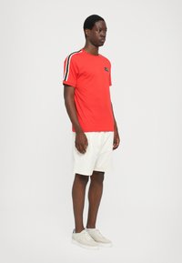 Red cotton t-shirt with black and white stripes on shoulders, paired with white shorts and light sneakers. Small logo detail on chest.