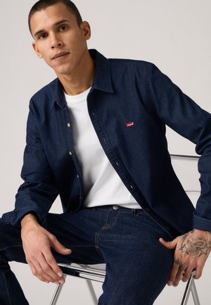 Camisa de botones de mezclilla oscura con un logo rojo, llevada sobre una camiseta blanca y combinada con jeans oscuros; presenta un cuello y un ajuste casual.