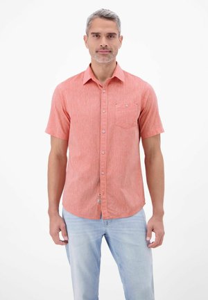 Mężczyzna stoi, ubrany w koralową koszulę na krótkim rękawie zapinaną na guziki oraz jasnoniebieskie dżinsy, na jednolitym białym tle.