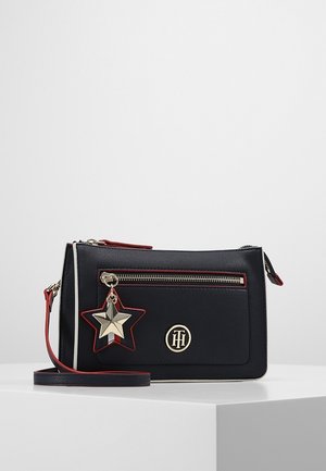 Sort crossbody-taske med lynlåslomme foran, guldfarvet stjernevedhæng, røde accenter og rundt guldfarvet logo på en hvid overflade.