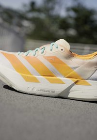 Lichtgekleurde hardloopschoen met oranje diagonale strepen, lichtblauwe veters en een dikke zool met het label "Lightstrike Pro" op asfalt.