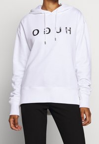 Sudadera blanca con capucha y "HUGO" impreso en negro, con cordones y puños acanalados, combinada con pantalones negros.