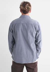 Camisa de pana azul con yugo trasero, mangas largas y dobladillo redondeado. Presenta una pequeña etiqueta con el logotipo cerca del cuello.