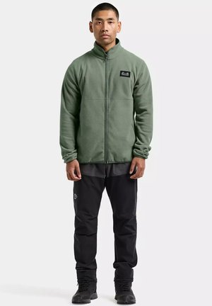 Man staat en draagt een olijfgroene fleecejas met rits, zwarte broek en zwarte laarzen tegen een effen lichte achtergrond.