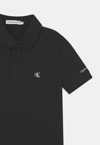 Camisa polo negra hecha de tejido texturizado, con una tapeta de tres botones. Presenta un logo blanco "cK" en el pecho y el nombre de la marca en la manga.