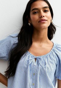 Lichtblauw gestreepte blouse met knopen aan de voorkant, korte pofmouwen en een strikdetail bij de halslijn. Glad materiaal met een verticaal patroon.