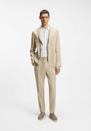 Man standing wearing beige suit with white textured shirt and light brown suede loafers against a plain white background.