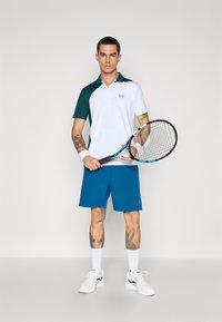 Polo de ténis em branco e verde escuro, com um logótipo. Combinado com calções azuis, meias brancas e sapatilhas brancas. A segurar uma raquete de ténis.