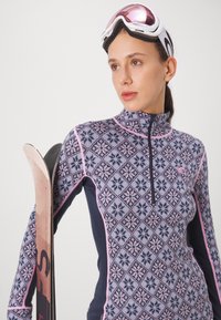Fleece skijakke med mønster i blått, rosa og marineblått. Har krage med glidelås og kontrastdetaljer, og er kombinert med et skistav.