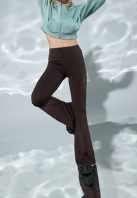 Pantalones acampanados marrones con una textura suave y elástica; combinados con una sudadera corta con cremallera en un suave tono azul claro. El fondo presenta sombras suaves y onduladas.