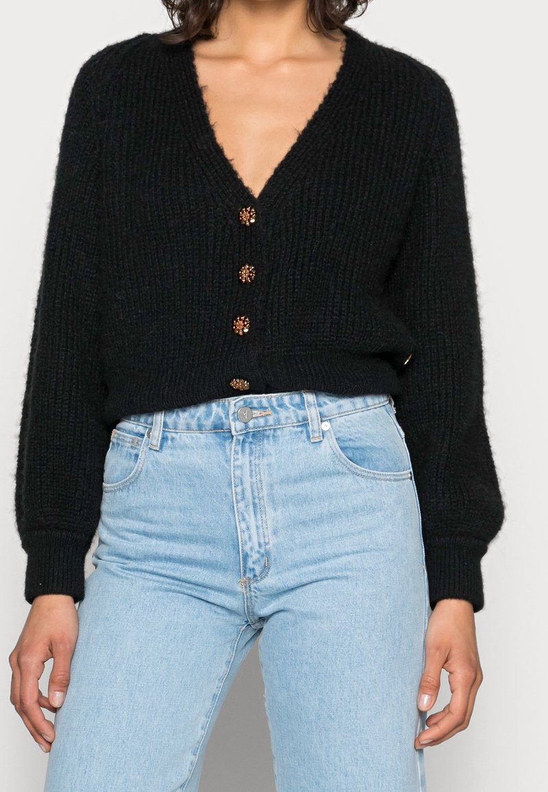 Femme portant un cardigan noir en maille à col en V avec des boutons dorés et un jean taille haute bleu clair, mains détendues le long du corps.