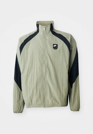 Veste zippée vert clair avec des accents bleu marine, manches raglan et tissu texturé ; présente un logo de marque sur le côté supérieur gauche.