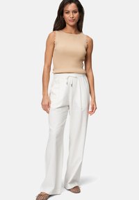 Débardeur beige à côtes avec des bords festonnés associé à un pantalon large blanc. Le modèle porte des chaussures à enfiler à motif avec des orteils ouverts.