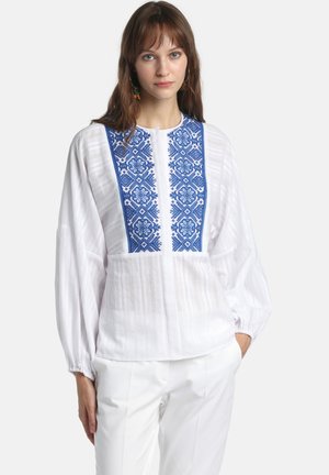 Nainen, joka pukeutunut valkoiseen puseroon, jossa on liehuvat hihat ja sininen geometrinen kirjailu rinnassa, yhdistettynä valkoisiin housuihin, kädet taskuissa.