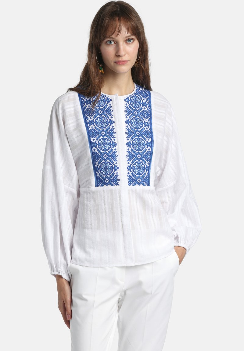 Femme portant un chemisier blanc à manches bouffantes avec une broderie géométrique bleue sur la poitrine, associé à un pantalon blanc, les mains dans les poches.