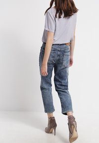 Kvinna som bär en ljusgrå t-shirt med kort ärm, blekta blå jeans och taupe högklackade ankelskor, stående med ryggen vänd mot kameran.