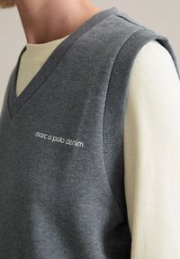 Grauer ärmelloser Pullover mit V-Ausschnitt und gerippten Kanten. Auf der Vorderseite befindet sich das weiße "marc o polo denim" Logo. Übereinander getragen mit einem cremefarbenen Hemd.