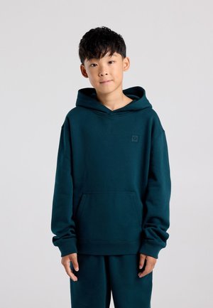 Garçon aux cheveux courts noirs portant un sweat à capuche bleu sarcelle foncé et un pantalon assorti, debout devant un fond uni gris clair.