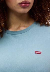 Κοντινό πλάνο του κάτω μέρους του προσώπου και του λαιμού ενός ατόμου που φοράει ανοιχτό μπλε πουκάμισο Levi’s με κόκκινο λογότυπο στο στήθος.