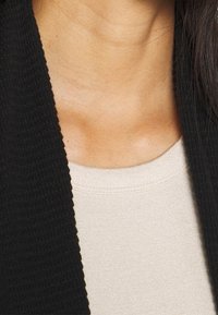 Schwarzer strukturierter Cardigan, der über einem hellbeigen Oberteil getragen wird. Der Stoff ist weich und zeigt sichtbare Nähte am Ausschnitt, die ein einfaches Design zur Schau stellen.