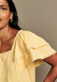 Blusa gialla in tessuto morbido, con scollatura quadrata, maniche a volant a strati e bordo in pizzo lungo i lati per un tocco di dettaglio in più.