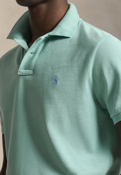 Άνδρας που φορά ένα ανοιχτοπράσινο πόλο πουκάμισο με ένα μπλε κεντημένο λογότυπο αλόγου και αναβάτη στο στήθος.