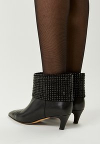 Botas de tobillo de cuero negro con un puño texturizado y tachonado, tacón delgado y punta afilada. El puño presenta un diseño de tachuelas geométricas.