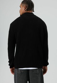 Zwarte gebreide trui met een hoge kraag, een gestructureerd ribpatroon en loszittende mouwen, gedragen over een wit overhemd en grijze broek.