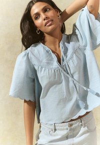 Light blue, short-sleeve blouse with a textured yoke, scalloped hem, and tie closure with tassels. Paired with light denim shorts.