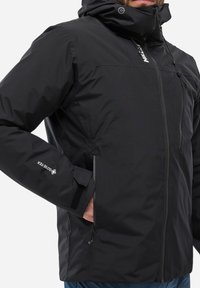 Schwarze, wasserdichte Jacke mit Reißverschluss, verstellbarem Kapuzen und GORE-TEX-Logo auf dem Ärmeln. Sie hat eine glatte, matte Textur.