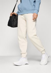 Cremefarbene Sweatpants mit schmal zulaufenden Beinen, versehen mit einem weißen Logo, kombiniert mit weißen Sportschuhen und einer schwarzen Tasche mit Netz-Akzenten. Hellblaues Hoodie getragen.