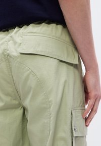 Τα cargo παντελόνια σε ανοιχτό πράσινο χρώμα έχουν υφή στην επιφάνεια, ελαστική μέση, πλαϊνές τσέπες και μια τσέπη με πατς με λεπτομέρεια κουμπιού.