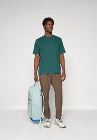 Mand iført en grøn kortærmet skjorte, brune bukser og hvide sneakers, der holder en lysblå rygsæk mod en ensfarvet baggrund.