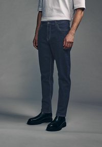 Jeans de mezclilla gris oscuro con un corte ajustado, con costuras en contraste y cierre frontal de botón, combinados con botas de suela de goma negras.