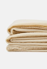 Vaalean beige kankainen peitto, joka on siististi taitettu kolmeen kerrokseen, ja siinä on pehmeä rakenne ja hienovarainen raidoitus reunoilla.