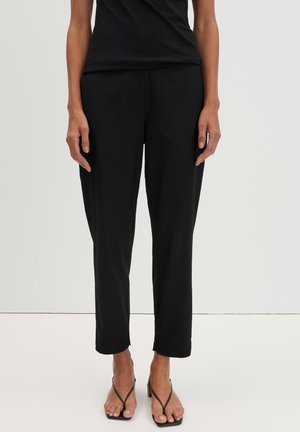 Femme debout portant un pantalon noir droit, un haut noir sans manches et des sandales noires à talons avec de fines lanières.