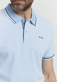 Svetlo modra polo majica iz teksturirane tkanine. Ima dvobarvno zapenjanje z gumbi in kontrastni ovratnik ter črtaste poudarke na rokavih.