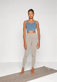 Mujer de pie descalza sobre una esterilla de yoga, vistiendo un top corto azul y leggings de talle alto beige, frente a un fondo liso.