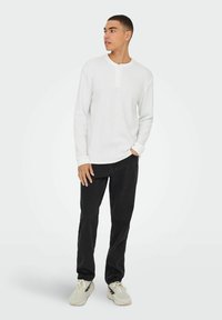 Weißes Langarm-Henley-Shirt aus strukturiertem Stoff, mit Knopfleiste, kombiniert mit schwarzen Jeans und hellgrauen Turnschuhen.