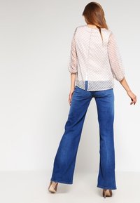 Blouse légère et transparente de couleur rose pâle avec des manches bouffantes, motif texturé ; associée à un jean flare bleu ; détail de fente au dos de la blouse. Des talons hauts viennent compléter le look.