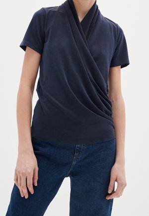 Persoon draagt een donker marineblauw kort mouw wikkelshirt gecombineerd met blauwe spijkerbroek, staand tegen een effen lichte achtergrond.