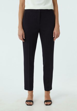 Femme portant un pantalon noir slim-fit jusqu'à la cheville, associé à des sandales noires à bout ouvert et un top blanc sans manches sur un fond uni.