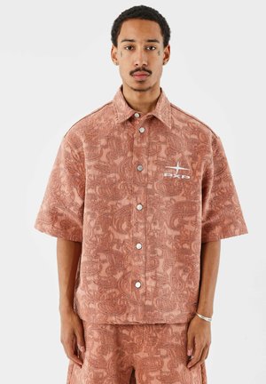 Man in een roestkleurig, gestructureerd overhemd met korte mouwen en bijpassende korte broek, staand tegen een witte achtergrond.