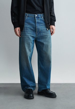 Uomo che indossa jeans blu larghi, giacca nera con zip, maglietta nera e scarpe nere grosse, in piedi su un pavimento grigio contro uno sfondo neutro.